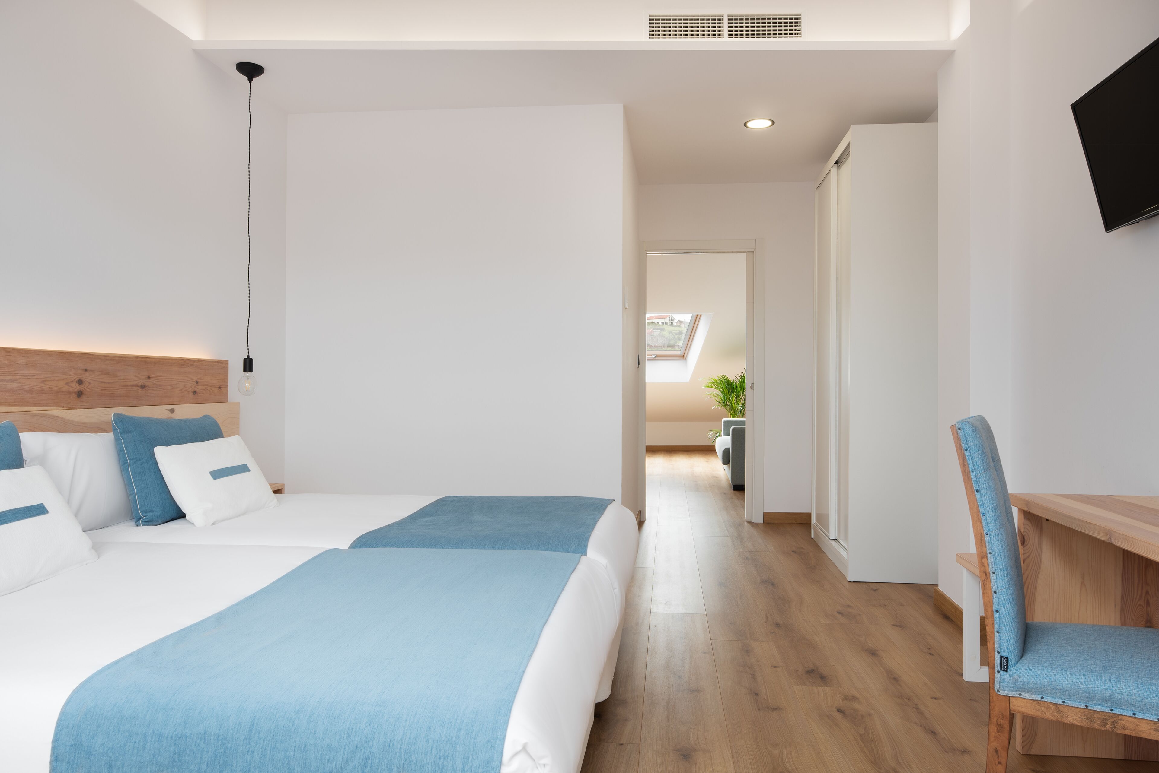 A modern hotel room with two beds, a wooden headboard, a desk with a chair, and a TV on the wall.