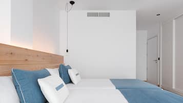 Un dormitori modern amb un capçal de llit de fusta, dos llits amb roba de llit blava i blanca, una lluminària penjant i una porta blanca.