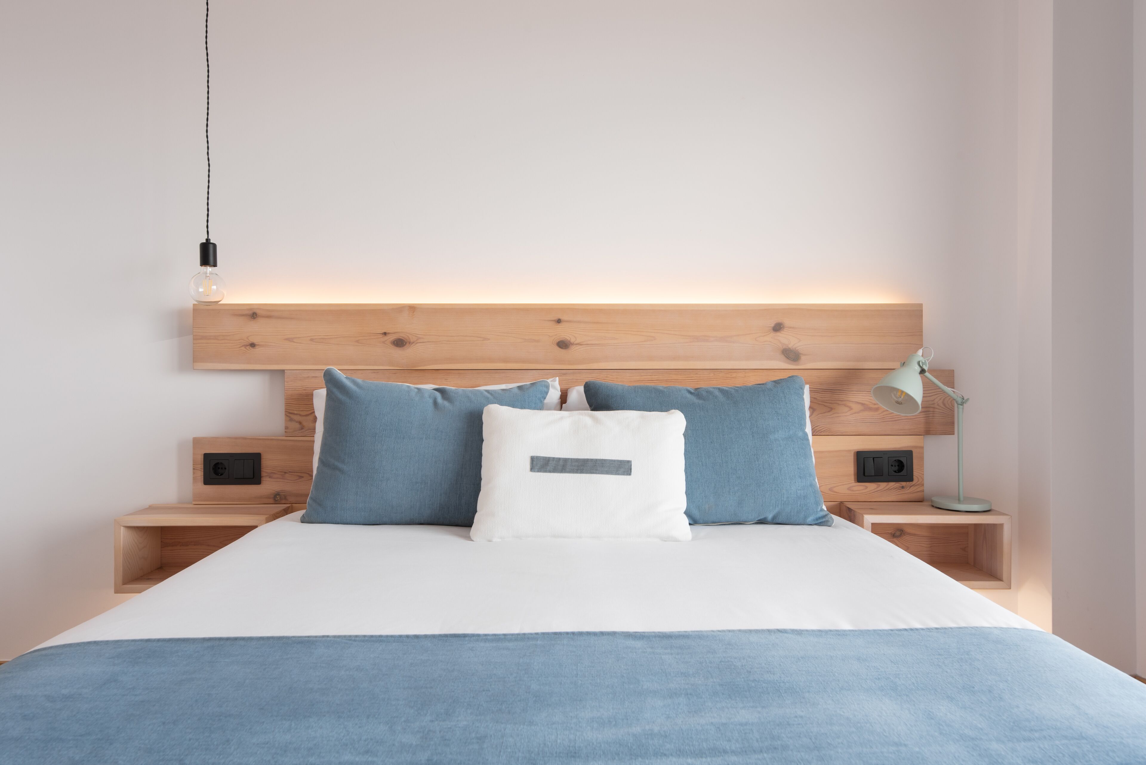A modern bedroom with a wooden headboard, a white bedspread, and two nightstands with a lamp and a power outlet.