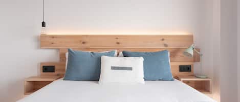 Une chambre à coucher moderne avec une tête de lit en bois, un couvre-lit blanc et deux tables de chevet équipées d’une lampe et d’une prise électrique.