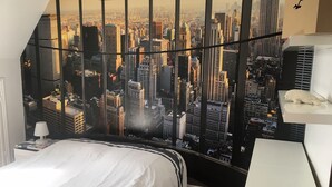 Un dormitorio con un mural de paisaje urbano en la pared, una cama con ropa de cama blanca, una mesita de noche y un pequeño estante con una lámpara.