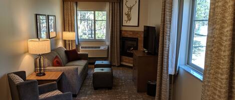 A hotel room with a sofa, armchair, ottoman, fireplace, television, and large windows with curtains.