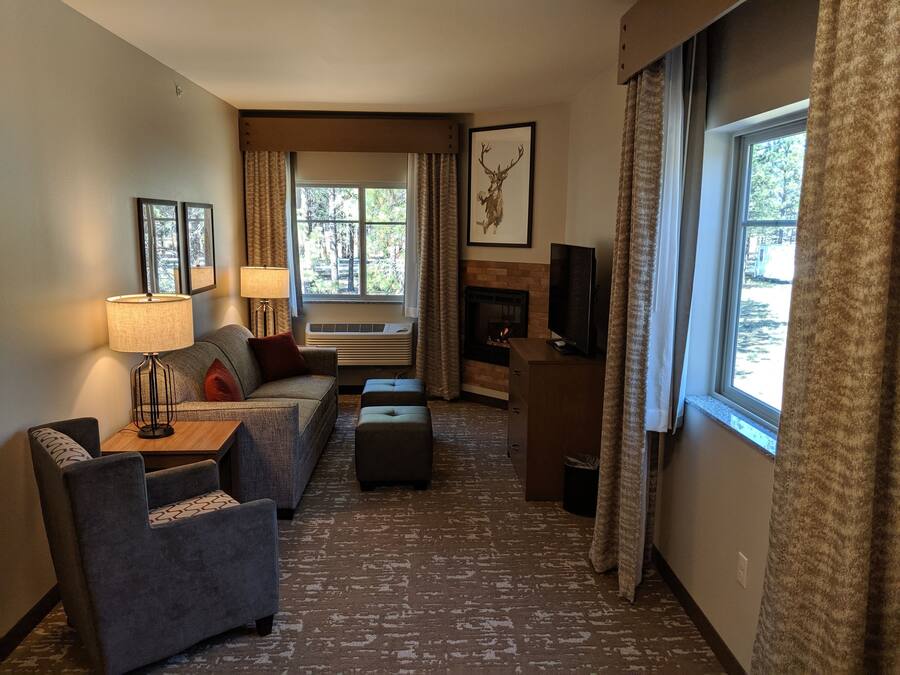 Habitación de hotel con sofá, sillón, reposapiés, chimenea, televisor y ventanas grandes con cortinas.