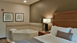 A hotel room with a large bed, a bathtub, a bedside table with a lamp, and two framed pictures on the wall. - GrandStay Hotel & Suites (Sisters)