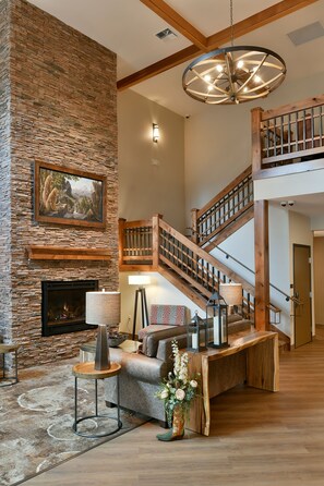 A spacious living area with a stone fireplace, a wooden staircase, and a large chandelier. - GrandStay Hotel & Suites (Sisters)