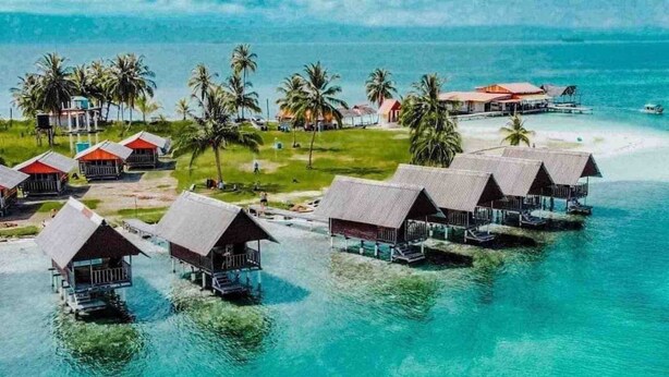 Overwater bungalows with thatched roofs, connected by walkways, and surrounded by clear turquoise water and palm trees. - Private Over the Water Cabin on San Blas Island (Las Huacas)