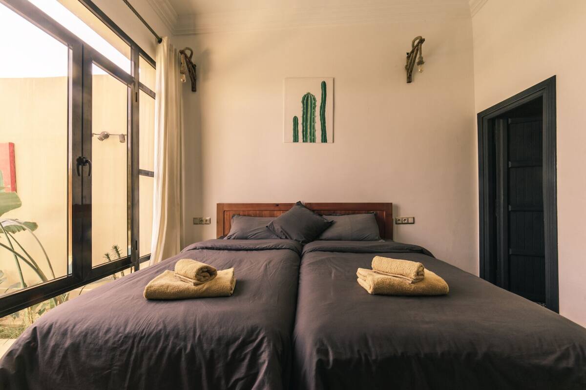 Ein Doppelbett mit dunkler Bettwäsche und zwei Handtüchern, ein Holz-Kopfteil, ein gerahmtes Bild einer Kaktee und ein Fenster mit Vorhängen.