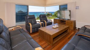 A modern living room with leather sofas, a wooden coffee table, and a television. Large windows offer a view of the ocean and a rocky landscape.