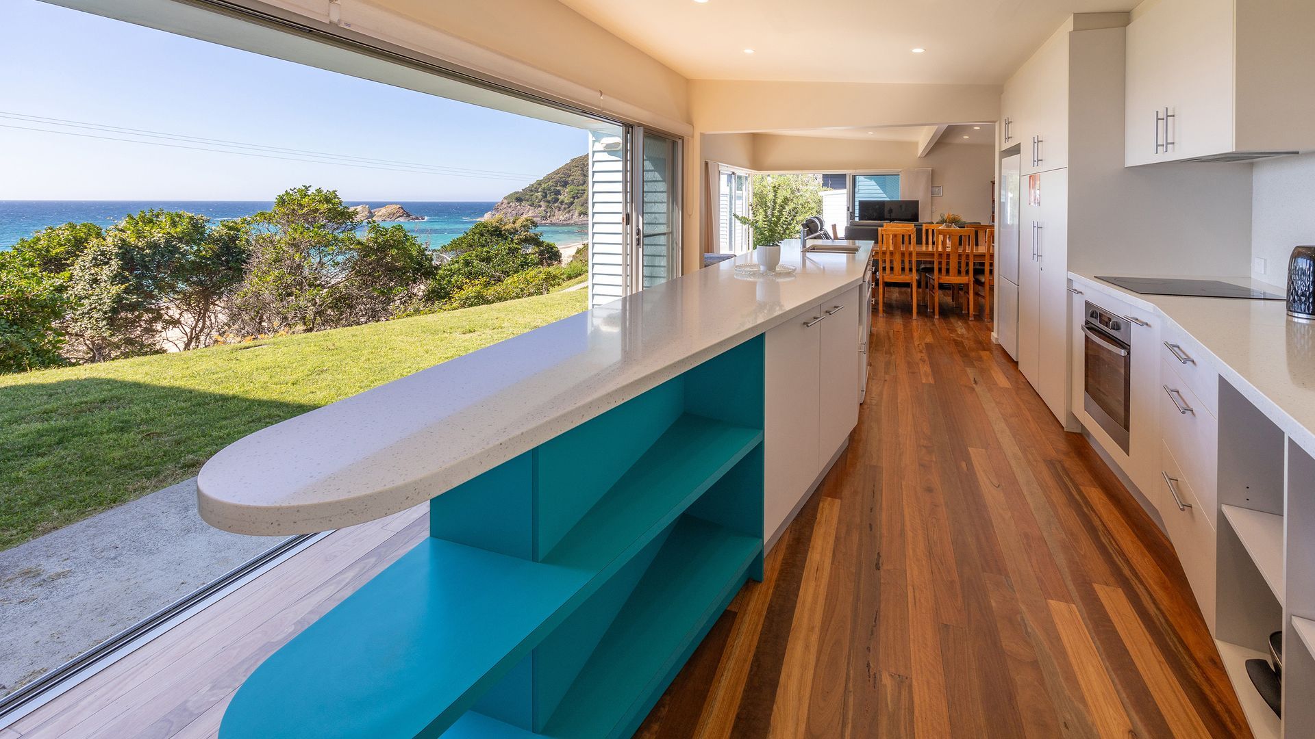 A modern kitchen with a view of the ocean, wooden flooring, and a dining area with chairs.