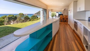 A modern kitchen with a view of the ocean, wooden flooring, and a dining area with chairs.
