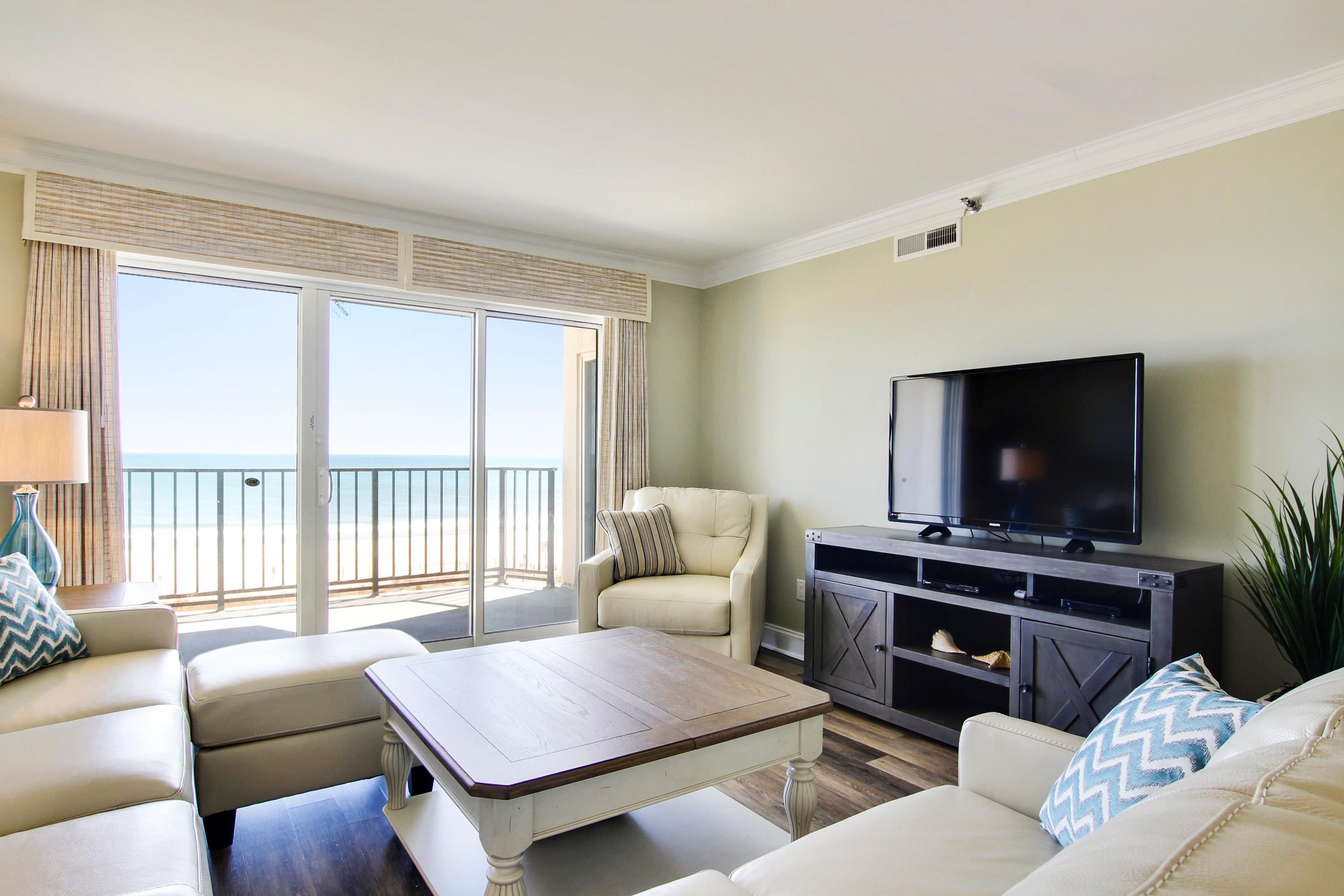 A living room with a television, a sofa, a coffee table, and a view of the beach through large windows.