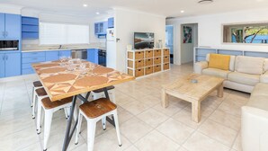 A modern kitchen with blue cabinets, a dining table with a geometric pattern, and a living area with a sofa and a coffee table. - Reflections - Pacific Palms Holidays (Smiths Lake)