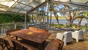 A covered outdoor seating area with a wooden table and chairs, overlooking a parking area and a body of water. - Reflections - Pacific Palms Holidays (Smiths Lake)