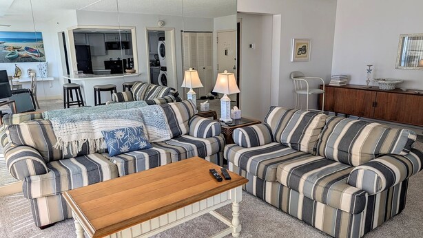A living room with striped sofas, a wooden coffee table, and a kitchen area in the background.