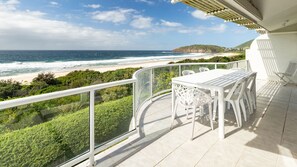 Un balcon avec une table et des chaises blanches donnant sur une plage et l’océan.
