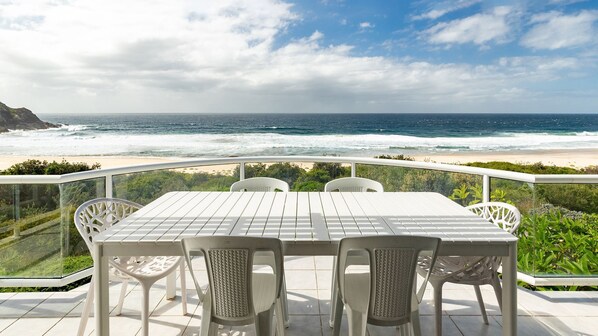 Une table et des chaises sur un balcon donnant sur une plage, avec des vagues et un ciel nuageux.