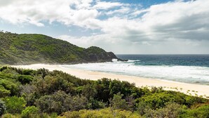 A coastal landscape with a sandy beach, lush green hills, and the ocean.