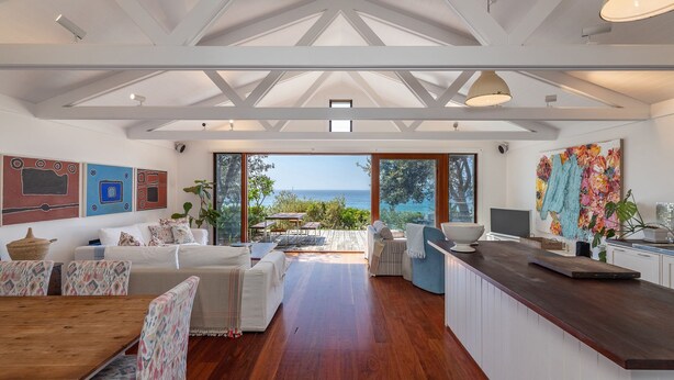A spacious living area with a wooden dining table, a sofa, and a kitchen counter. There are large windows offering a view of the sea. - Barefoot on Blueys - Blueys Beach (Blueys Beach)
