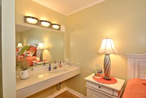 A bathroom with a large mirror, two wall-mounted lights, a sink, a vanity, and a bedside table with a lamp.