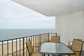A balcony with a glass table and wicker chairs, overlooking a beach and the ocean.