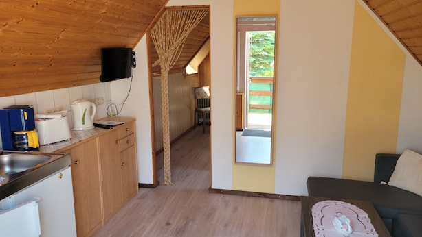 A compact kitchen with wooden cabinets, a coffee maker, and a toaster. A small dining area with a table and a decorative item is visible. - Apartment morning sun (Pruchten)