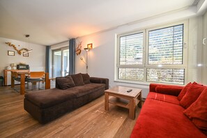 Una sala de estar con un sofá marrón, un sofá rojo, una mesa de centro de madera, un comedor con mesa y sillas, y un ventanal con persianas.