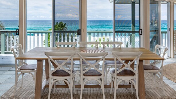A dining area with a wooden table and white chairs, overlooking a turquoise ocean through large glass doors.