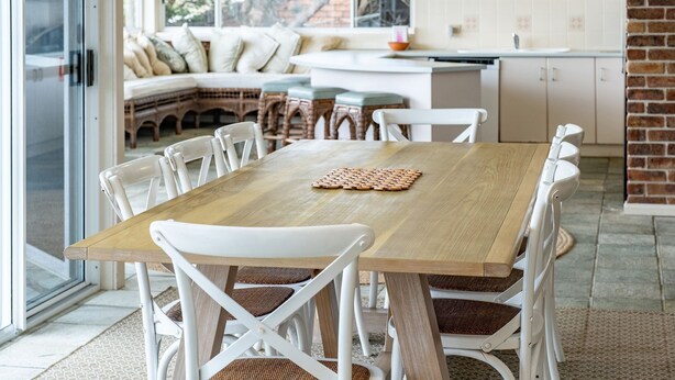 A dining area with a wooden table and white chairs, a woven placemat, and a kitchen in the background.