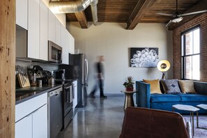 A modern kitchen with stainless steel appliances, a dining area with a blue sofa and a small table, and a large window providing natural light.