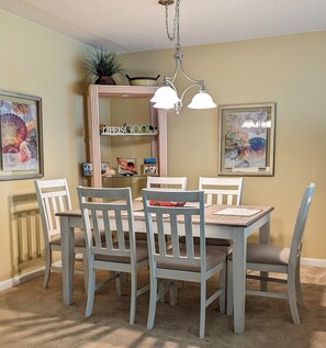 A dining room with a wooden table and chairs, a chandelier, framed artwork, and a shelf with decorative items.