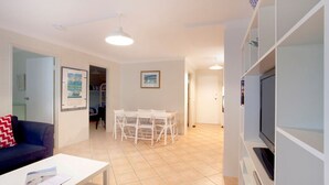A living room with a sofa, a dining table with chairs, a TV, and a kitchen area with cabinets. - Blueys Beach Villa Manyana 24 (Blueys Beach)