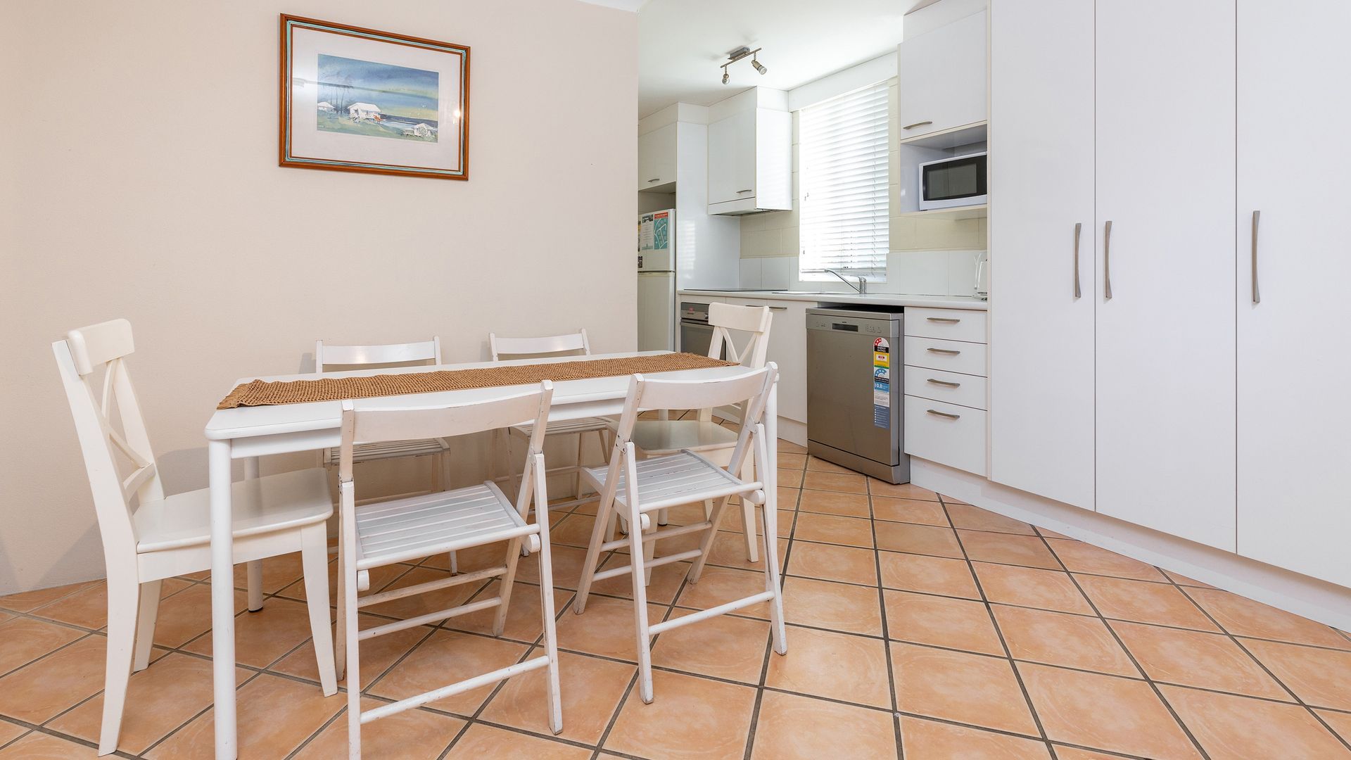 A dining area with a white table and chairs, a kitchen with white cabinets and a microwave, and a tiled floor.