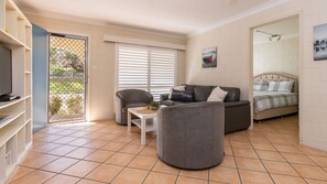 Un espace de vie compact comprenant un canapé, un fauteuil et un lit. On y trouve un téléviseur posé sur un meuble, une petite table avec une plante et une fenêtre avec des stores.