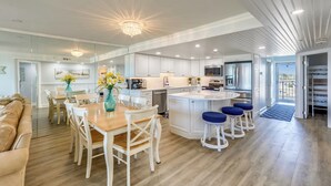 A modern kitchen with white cabinetry, a central island with stools, and a dining area with a wooden table and chairs. - DAILY ACTIVITIES & LINENS INCLUDED*! Direct oceanfront 2BR/2BA with enclosed den (used as 3rd bedroom). (Ocean City)