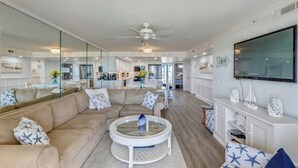 A modern living room with a large sofa, a round coffee table, and a television mounted on the wall. - DAILY ACTIVITIES & LINENS INCLUDED*! Direct oceanfront 2BR/2BA with enclosed den (used as 3rd bedroom). (Ocean City)