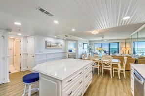 Cozinha moderna com armários brancos, bancada de mármore e área de refeições com cadeiras e mesa de madeira.