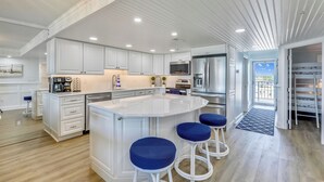 A modern kitchen with white cabinetry, a central island with stools, stainless steel appliances, and a view to the outside. - DAILY ACTIVITIES & LINENS INCLUDED*! Direct oceanfront 2BR/2BA with enclosed den (used as 3rd bedroom). (Ocean City)