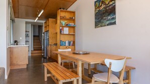 A modern kitchen with wooden cabinets, a dining table, and a bench. There is a bookshelf with books and a painting on the wall. - Sea Shack - at Seal Rocks (Seal Rocks)