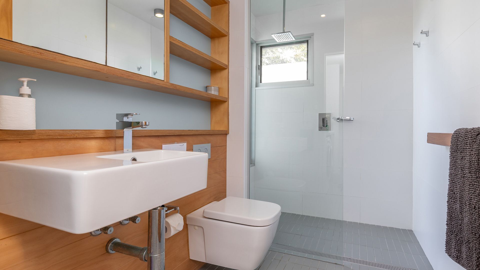A modern bathroom with a white sink, wooden shelves, a toilet, and a shower area.