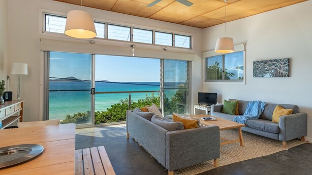 A modern living room with a large sliding glass door leading to a balcony with a view of the sea. There is a grey sofa, a wooden coffee table, and a TV stand with a flat-screen TV. - Sea Shack - at Seal Rocks (Seal Rocks)