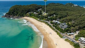 An aerial view of a coastal area with a sandy beach, a road, and buildings surrounded by greenery.