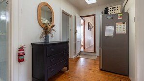 A hallway with a wooden floor, a black dresser with drawers, a mirror, a fire extinguisher, and a refrigerator with papers on it.