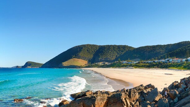A coastal beach with rocky foreground, clear blue sea, and houses on a hillside in the background. - Banksia - Pacific Palms Holidays (Blueys Beach)