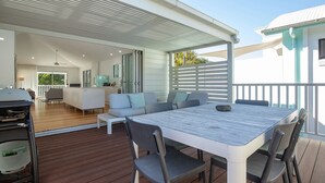 A modern outdoor dining area with a wooden deck, a white table, and grey chairs. - Banksia - Pacific Palms Holidays (Blueys Beach)