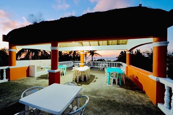 Un espace extérieur aménagé en terrasse, avec des tables et des chaises, un coin salon abrité et une rambarde blanche.