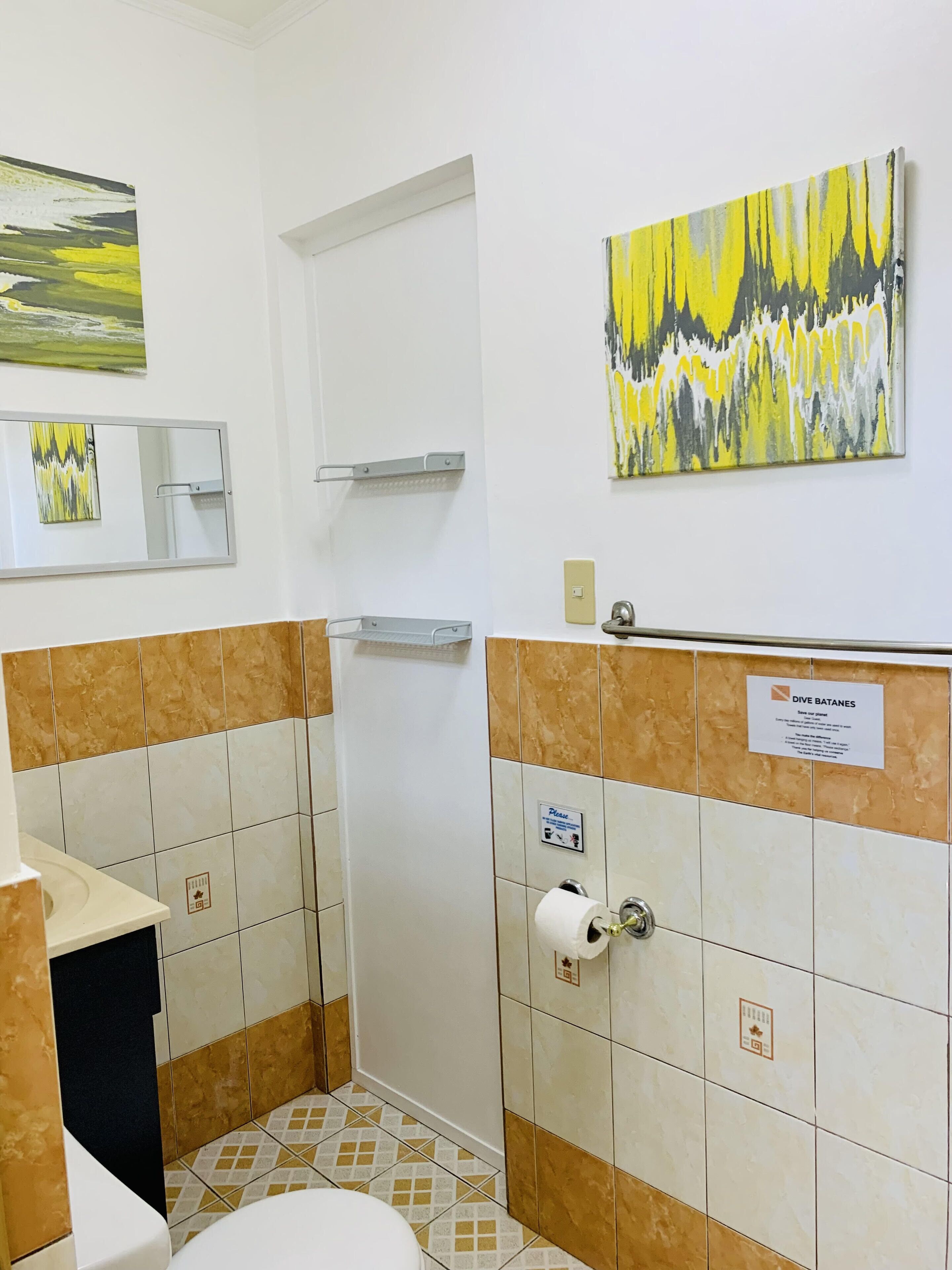 A bathroom with a toilet, sink, and shower. There are two paintings on the wall, a sign above the sink, and a towel rack.