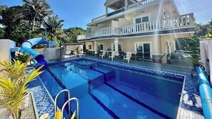 Una piscina con tobogán, un edificio de dos pisos con balcón y palmeras al fondo.