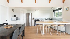 A modern kitchen with white cabinetry, a stainless steel refrigerator, a wooden dining table with bar stools, and a window providing natural light. - Ferny Lane House (Norfolk Island)