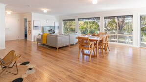 Uma sala ampla com sofá, mesa de jantar e cadeiras, janelas grandes com vista para árvores e piso de madeira.