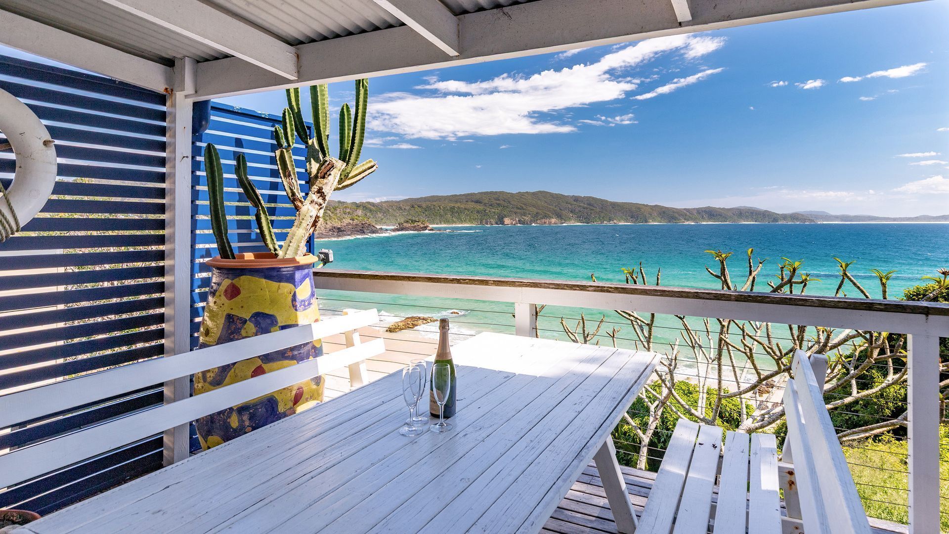 A wooden deck with a table, a potted cactus, and a bottle of champagne.